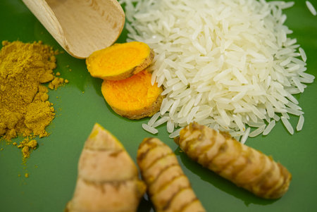 Fresh raw turmeric roots and slices next to heaps of rice and spice powder on a green plate. Selective focus and free space for textの写真素材