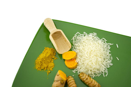 Fresh raw turmeric roots and slices next to heaps of rice and spice powder on a green plate with free space for textの写真素材