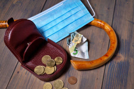 Conceptual  flat lay still life with money spilling from an open purse onto a wooden table with cane and surgical mask alongside viewed from aboveの写真素材