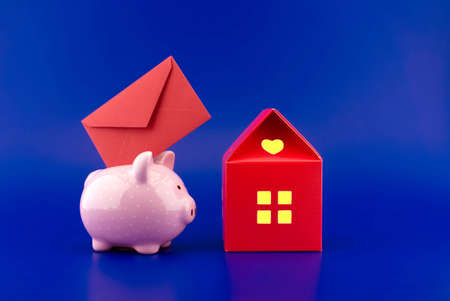Festive red envelope being inserted into a pink piggy bank next to a small red house of paper with warm light in windows over blue background with copyspaceの写真素材