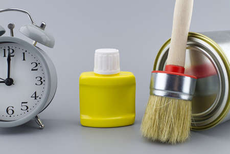 DIY, decorating or renovations concept with a still life of an alarm clock with assorted clean paint brushes and unlabeled tin of paint over a grey backgroundの写真素材