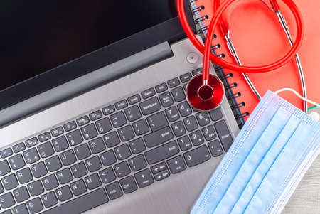 Virus pandemic and medical concept with a colorful red stethoscope coiled over a surgical face mask alongside a notebook on a wooden desk with computer keyboard viewed top downの写真素材