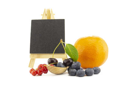 Fresh seasonal fruit still life with small chalkboard and assorted berries including blueberries, cherries and red currants on wooden spoons and orange on a white backgroundの写真素材