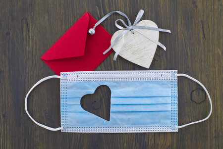 Face mask with cut out heart shape and red envelope on wooden background with free copy space. Social distancing and contact-less greetingsの写真素材