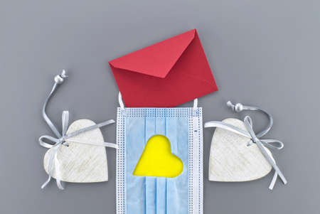 Two hearts between a medical face mask with cut out heart shape and red envelope on a gray background. Social distancing and contact-less greetingsの写真素材