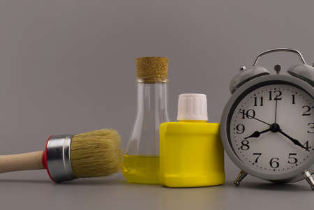 DIY, decorating or renovations concept with a still life of an alarm clock with clean paint brush, yellow pigment container and laboratory flask over a grey background with free space for textの写真素材
