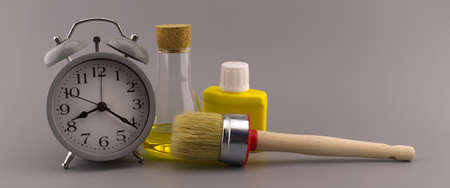 New color, decorating or renovations concept with a still life of an alarm clock with clean paint brush, unlabeled yellow pigment container and laboratory flask over a grey backgroundの写真素材