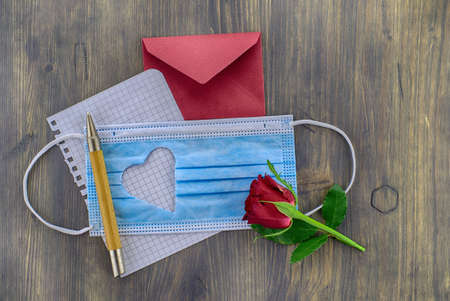 Face mask with cut out heart shape alongside a wire bound notepad and pen and red envelope on wooden background with free copy space. Social distancing and contact-less greetingsの写真素材