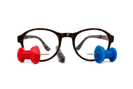 Reading glasses with tortoiseshell frames, pen and colorful red and blue thumb tack over a white background with copy space in a business still lifeの写真素材