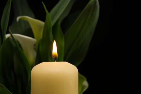 Close up of candle burning flame in the darkness and arum lilies illuminated by the candlelight alongside in a conceptual imageの写真素材