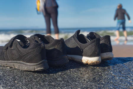 Ground level view of two pairs of casual black shoes on a rock at the beach with blurred figures and the ocean in the background in a summer vacation conceptの写真素材