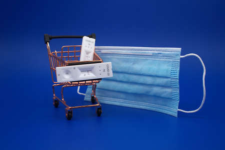 Covid-19 and coronavirus healthcare and shopping concept with mask, shopping cart and rapid antibody test kits over a blue backgroundの写真素材