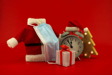 Christmas in pandemic concept still life with vintage alarm-clock, miniature santa costume and face mask, gift boxes on a festive red backgroundの写真素材