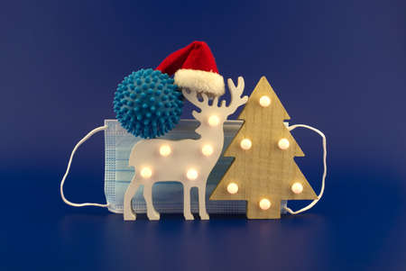 Christmas in pandemic concept still life with blue virus molecule, red Santa hat, Christmas tree, gift box and surgical face mask all isolated on blue backgroundの写真素材