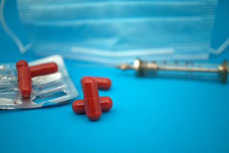 Medical, Covid-19 and healthcare background with red capsules scattered on blue alongside an empty blister pack with syringe and face maskの写真素材