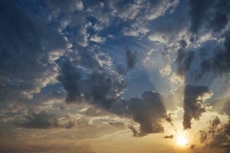 Stunning orange and blue sky with a setting sun behind a layer of fluffy white clouds, casting a beautiful backlighting effectの写真素材