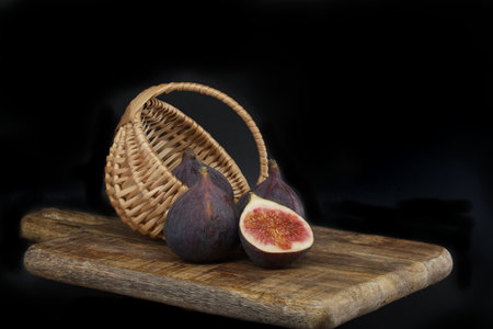 Figs whole and sliced in half near wicker basket on a rustic wooden board over dark background