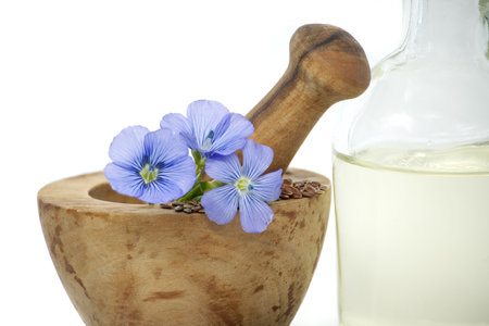 Wooden mortar filled with small brown flax seeds and a blue flax plant flowers alongside linseed oil in glass decanter isolated on white backgroundの写真素材