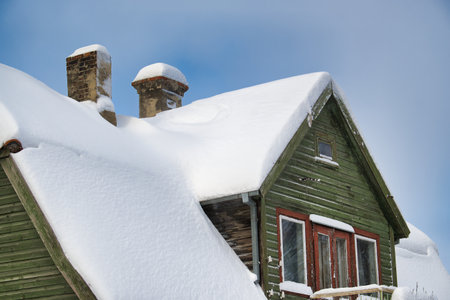 Serene winter scene focused on a green wooden house. The house's roof is heavily covered in white snow, with two chimneys protruding from the thick layer of snowの写真素材