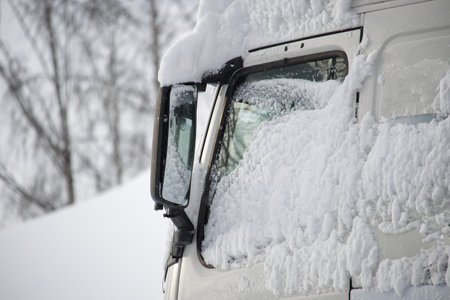 Close-up of a white truck windshield, hood, and side mirror are notably covered in thick, fluffy snow, vehicle is partially buried under the snowの写真素材