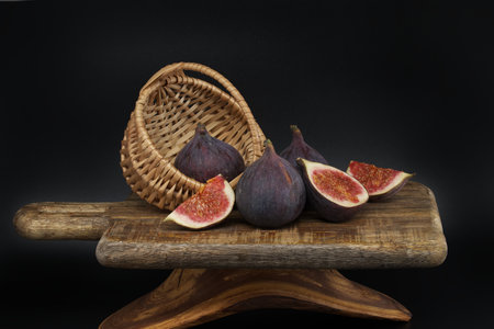 Figs whole and sliced in half near wicker basket on a dark wooden boardの写真素材