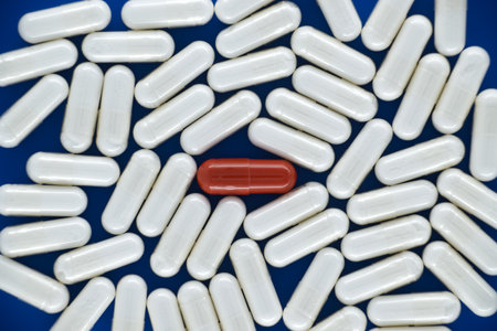 Numerous white pills are spread out over a blue background with one red pill situated in the center, drawing attention from its white capsulesの写真素材