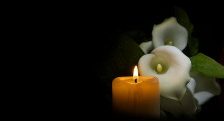 Single burning wax candle radiating a gentle flame next to white calla lily flower in a dark environment. Close-up view with free copy spaceの写真素材