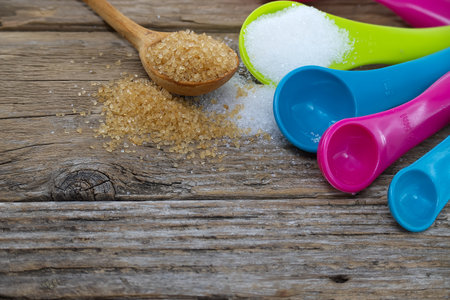 Set of colorful plastic measuring spoons with brown cane sugar and white sugar spilled around on a rustic wooden surfaceの写真素材