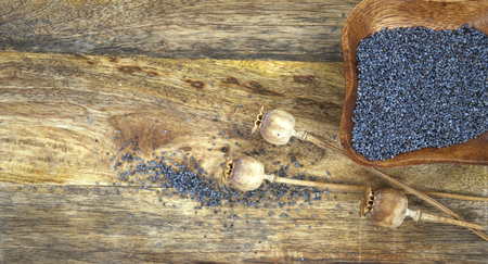 Small wooden bowl filled with black poppy seeds and dried poppy seed heads on rustic wooden tableの写真素材
