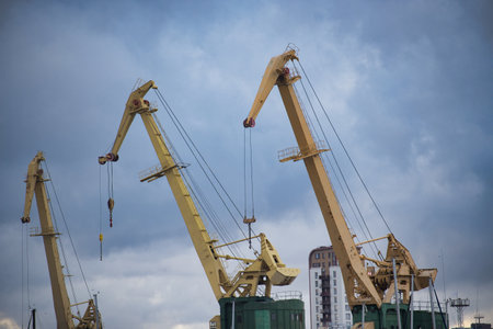 Three large industrial cranes in a harbor, showcasing construction and shipping industry elements under a dramatic sky.の写真素材