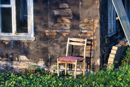 Old, decaying house wall with a rustic wooden chair surrounded by lush greenery, capturing a sense of abandonment and nostalgia in the sunlight.の写真素材