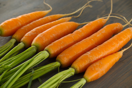 A close-up of fresh orange carrots with vibrant green stems placed on a wooden background, showcasing their natural and healthy appeal.の写真素材
