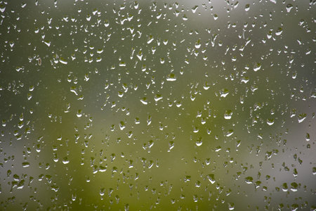 Raindrops on a window glass creating a soft focus for the greenery outdoors, evoking serenity and...