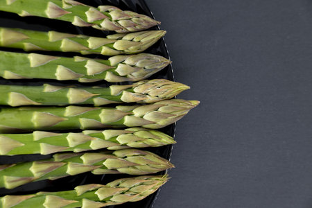 Fresh green asparagus spears arranged on a black plate against a dark gray background, highlighting the vibrant vegetables and textures.の写真素材