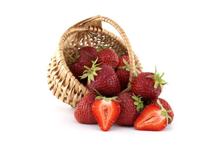 Ripe, juicy strawberries are spilling out of a small wicker basket onto a clean white background, with one strawberry sliced in half, showcasing the vibrant red interior and freshness of the fruitの写真素材
