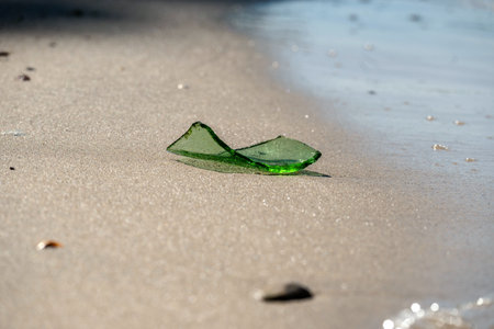 A single green glass fragment rests on soft sandy beach, facing the gentle waves. Captures contrast...