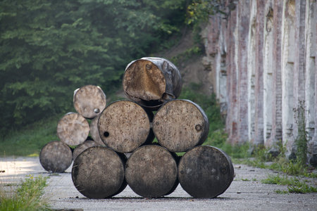Weathered metal barrels stacked outdoors near an old brick wall surrounded by lush forest greenery. The rustic and abandoned atmosphere creates a contrast between nature's growth and industrial decay.の写真素材
