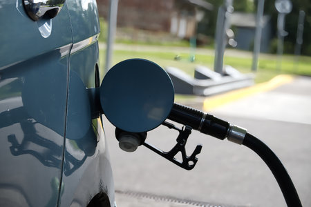 Close-up of a fuel pump nozzle inserted into a blue car's tank under bright daylight. The scene...