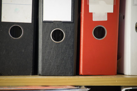 A close-up of office binders in various colors. The binders are neatly arranged on a wooden shelf,...