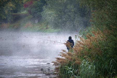 A person stands on the bank of a tranquil lake, fishing in the early morning mist. The scene is framed by colorful trees and reeds.の写真素材