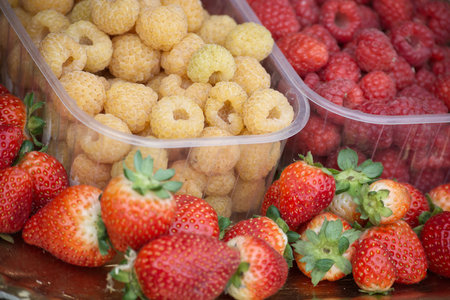 Bright market display of ripe strawberries, red raspberries, and yellow raspberries in clear plastic containers. Ideal for topics like fresh produce, healthy snacking, and colorful fruitの写真素材
