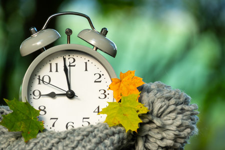 A close-up of an analog clock, surrounded by colorful fall foliage and a soft, gray scarf, evoking a...