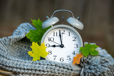 A classic alarm clock is surrounded by colorful autumn leaves and a cozy knitted scarf, evoking the season's warmth.の写真素材