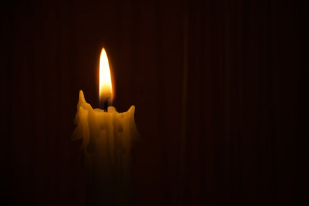 Solitary candle burns in dark setting, bright flame casting a soft, amber light. The melting wax trails down the sides, creating a serene, intimate atmosphere suitable for reflection and calm momentsの写真素材