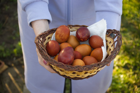 A person holds a wicker basket overflowing with a collection of beautifully dyed Easter eggs. The...
