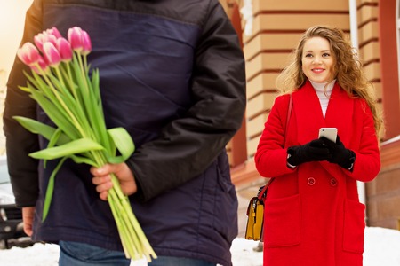 Romantic date. Man hides the flowers behind his back for his girlfriend. Beautiful young couple walking together through city streets. Spring.の写真素材
