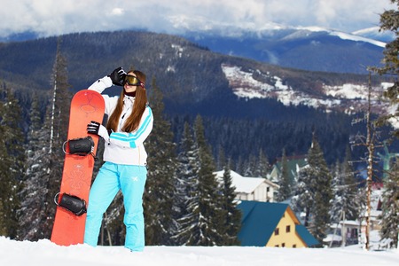 Young beautiful smiling girl in white jacket, blue ski pants and googles on her head standing with snowboard in the snowy mountains. Winter sports.の写真素材