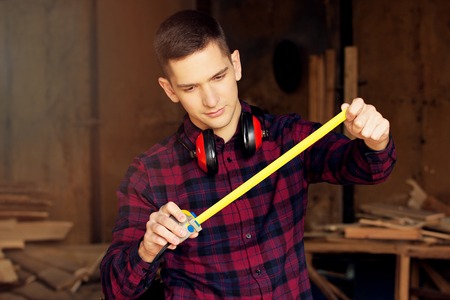 Workman dressed in the checkered shirt working with tape-measure at the sawmill. Timbers on background.の写真素材
