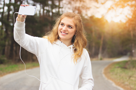 Sports selfie. Portrait of young and attractive redhaired sports girl taking a selfie on her phone and listening music in the park.の写真素材