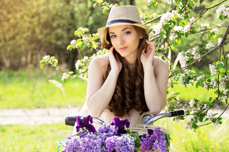 Portrait of young beautiful girl in hat with long hair with flowers in basket on vintage bike. Fashioned woman.の写真素材